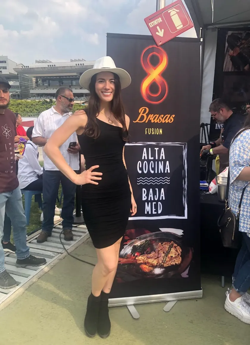 Mujer con sombrero y vestido negro posando frente a stand de alta cocina en evento de la agencia.