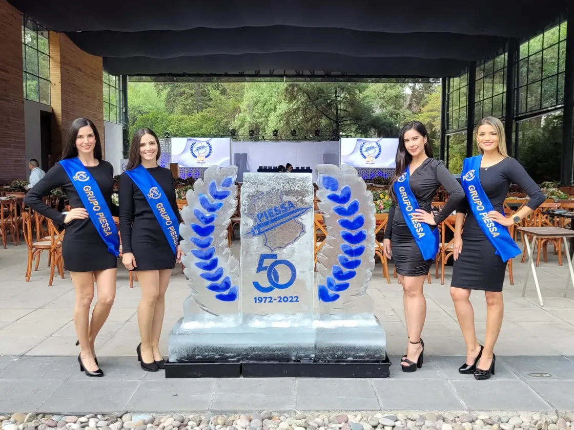 Grupo de edecanes con vestidos y bandas azules posando junto a escultura de hielo conmemorativa de la agencia.